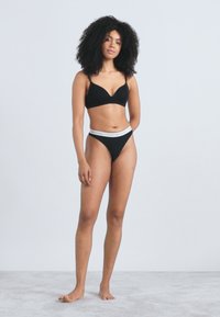 Črni komplet modrčka in tangic iz mehke tkanine. Modrček ima izrez v obliki črke V; tangice imajo bel rob z brandiranim logotipom.