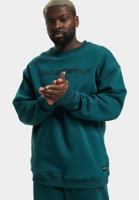 Teal színű pulóver ribbelt mandzsettákkal és szegéllyel, a mellkasán fekete "ROCAWEAR" logóval. Puha pamut keverékből készült.