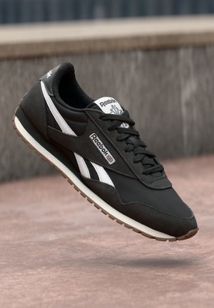 Zwarte Reebok sneaker met witte zijstrepen, gumzool en gebrande tong, zwevend boven een betonnen oppervlak met een vage achtergrond.