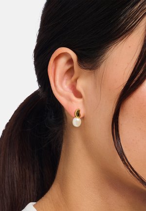 Goldener Steckerohrring mit einzelnen weißen Perltropfen, getragen am linken Ohr einer Person mit zurückgebundenem dunkelbraunem Haar.