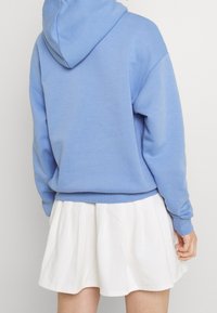 Osoba v světle modré mikině s kapucí a bílé plissé sukni, stojící zády k plochému světlému pozadí.