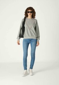 Sweatshirt cinza com um padrão decorativo em forma de V, combinado com calças skinny azuis e ténis brancos. O modelo está a segurar uma mala preta.