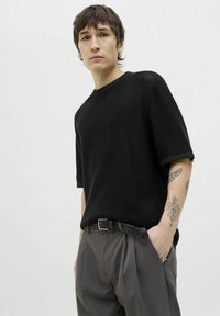Black knitted short-sleeve top with a textured weave, paired with gray pleated trousers and a braided belt, worn by a model facing forward.