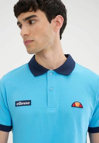 Polo shirt blu chiaro con colletto blu navy, dotato di tre bottoni e un patch con logo ricamato e dettaglio sul petto.