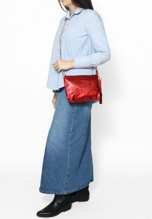 Borsa a tracolla rossa in metallo con superficie texturizzata, chiusura con zip e tracolla a catena, mostrata su una camicia azzurra chiara e una gonna di jeans.