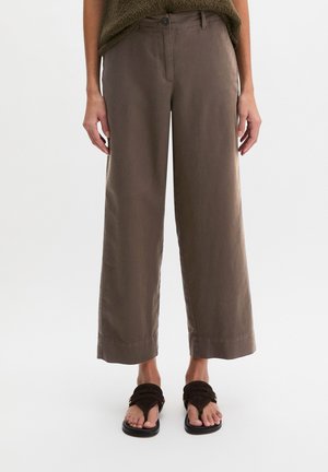 Bruine wijduitlopende cropped broek gedragen met zwarte sandalen en een groen gebreid topje, vanaf de helft van de romp tot aan de voeten getoond tegen een neutrale achtergrond.