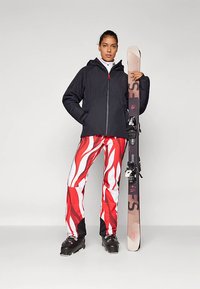 Schwarze isolierte Jacke mit Kapuze, lebendige rote und weiße Skihose mit Flammenmuster, schwarze Skischuhe und ein Ski mit Bindungen.
