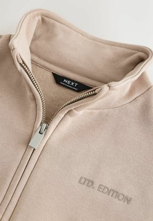 Sudadera beige con cremallera hecha de tela suave, con un alto cuello, cremallera plateada y el texto en relieve "LTD. EDITION" en el frente.