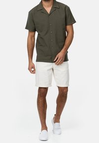Camisa de manga corta de color verde oliva con un cuello relajado, cierre frontal con botones, combinada con pantalones cortos blancos arremangados y zapatos blancos sin cordones.