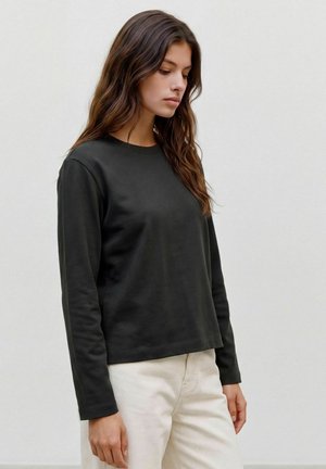 Jeune femme aux longs cheveux bruns portant un t-shirt noir uni à manches longues et un pantalon crème clair, debout devant un fond clair uni.