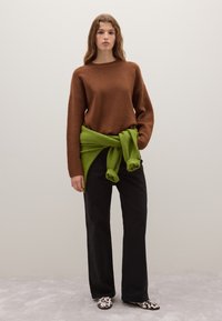 Maglione marrone a maglia, cintura verde in vita, pantaloni neri a gamba larga e scarpe a motivo bianco e nero. Dettagli del tessuto texture visibili.