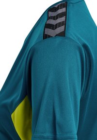 Close-up van een teal sportshirt schouder met zwart-wit chevron-patroon op de schoudernaad en een geel armsgedeelte.
