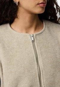 Vrouw met een beige, gestructureerde ritsvest, kleine zilveren ringoorbellen zichtbaar, met krullend donker haar en een neutrale lipkleur.