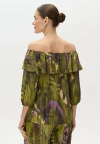Blusa off-the-shoulder de tejido ligero con un estampado de hojas en verde, morado y beige. Presenta un escote con volantes y mangas de tres cuartos.