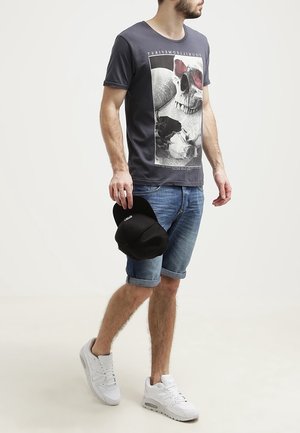 T-shirt grise avec un graphic de crâne, manches courtes et encolure ronde. Porté avec des shorts en denim retroussés et des baskets blanches, tenant une casquette noire.