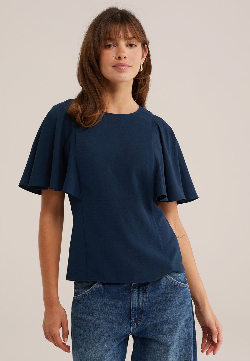 Femme portant un haut bleu marine avec de larges manches volantes et un jean bleu, debout devant un fond beige uni.