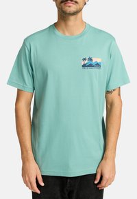 Mintgroene katoenen t-shirt met een graphic op de linkerkant van de borst, waarop palmbomen, bergen en een zonsondergang in blauw, roze en beige tinten zijn afgebeeld. Relaxed fit.