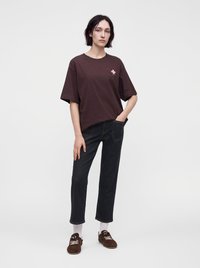 Άτομο που στέκεται, φοράει ένα φαρδύ καφέ t-shirt με μικρό λουλουδάτο σχέδιο, μαύρα κοντά τζιν, λευκές κάλτσες και καφέ παπούτσια.