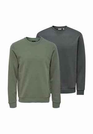 Junger Mann mit lockigem Haar, der einen olivgrünen Pullover, schwarze Hosen und braune Schuhe trägt, steht und schaut nach links.