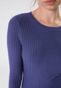 Suéter roxo de malha ribada com mangas longas e decote arredondado. O tecido é suave, com linhas verticais visíveis. Sem padrões ou detalhes.