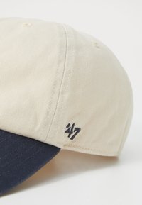 Baseballpet met een lichtbeige kroon en een marineblauwe klep, gemaakt van gestructureerde stof, met '47' geborduurd aan de zijkant.