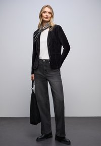 Femme blonde portant un blazer en velours côtelé noir, une chemise blanche, un jean large foncé, une écharpe imprimée léopard, des bottes noires, tenant un sac fourre-tout noir.