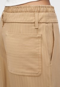 Κοντινή εικόνα μπεζ παντελονιού με λεπτές ρίγες, ελαστική μέση, θηλιές για ζώνη, μεγάλη ορθογώνια πίσω τσέπη και πλαϊνή τσέπη στη ραφή.