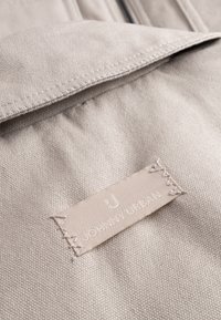 Beżowa tkanina o teksturowanym splocie, z naszytą etykietą, na której widnieje napis "JOHNNY URBAN" w jasnym kolorze nitki.