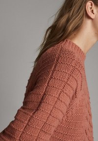Dzianinowy sweter w kolorze koralowym z teksturowanym, wzorzystym wzorem. Posiada wysoki dekolt oraz długie rękawy, prezentując misternie wykonane detale.