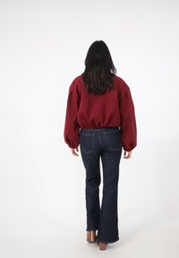Bordowa bluza z balonowymi rękawami i elastycznym dołem, zestawiona z ciemnoniebieskimi jeansami o rozszerzanych nogawkach. Osoba stoi tyłem na białym tle.