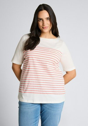 Femme aux longs cheveux foncés, portant une chemise blanche à rayures horizontales rouges et un jean bleu clair, debout les mains dans le dos.