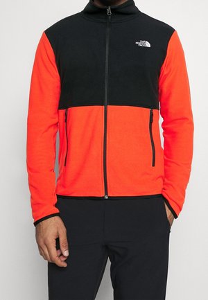 Mann trägt eine schwarze und leuchtend orangefarbene Fleecejacke mit Reißverschluss, seitlichen Reißverschlusstaschen und dem The North Face-Logo auf der Brust.