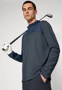Navy long-sleeve golf shirt with a lighter navy chest panel, a zipper collar, and smooth texture. Model holds a golf club, wearing a white glove.