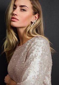 Top beige à sequins à manches courtes, orné de sequins variés et d'un col rond. De grandes boucles d'oreilles en cristal ajoutent une touche décorative.