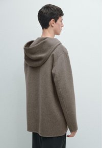 Indumento marrone con cappuccio realizzato in tessuto morbido e strutturato con una vestibilità rilassata. La parte posteriore presenta un collo alto e spalle scese.