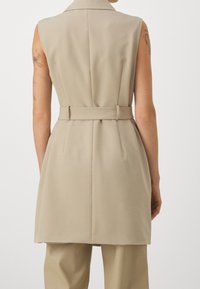 Jasnobeżowa bezrękawnikowa kamizelka o strukturalnym kroju, z paskiem w talii i przeszyciami z tyłu. Dopasowany materiał o gładkiej fakturze.