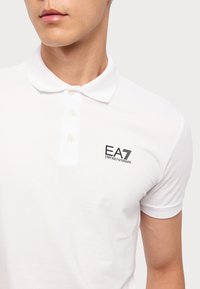 Hvit polo skjorte laget av bomull, med klassisk krage, tre knapper, og en brodert logo på venstre bryst. Korte ermer.