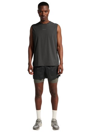 Athletischer Mann stehend, trägt ein ärmelloses schwarzes Oberteil, schwarze geschichtete Shorts mit grauer Innenschicht, weiße Socken und graue Laufschuhe.