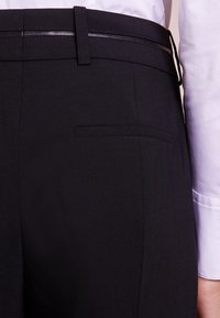Pantalons noirs en tissu texturé, avec une ceinture lisse et brillante et une poche arrière unique sans rabat.