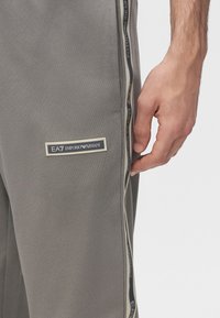Κοντινό πλάνο γκρι παντελονιού Emporio Armani με λωρίδα λογότυπου στο πλάι και ορατό χέρι που ακουμπά δίπλα στο πόδι.