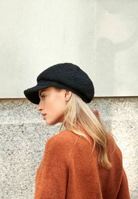 Zwarte, textuurvolle wollen hoed met een brede, rechte rand, gedragen onder een hoek. Het model heeft lang blond haar en draagt een oranje jas.