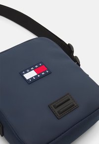 Granatowa torebka crossbody o gładkiej teksturze; zawiera prostokątny logo Tommy Jeans oraz czarną naszywkę. Dołączony regulowany pasek.