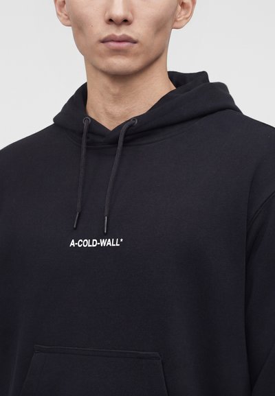 Άνδρας φορώντας μαύρο φούτερ με κορδόνια και άσπρο κείμενο "A-COLD-WALL*" στο στήθος, ουδέτερο γκρι φόντο.