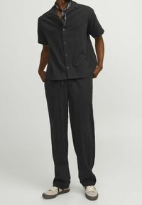 Mustat lyhythihaiset paidat, joissa on napit, yhdistettynä löysiin mustiin housuihin. Kaulassa on mustavalkoinen kuviollinen huivi. Rentoja jalkineita näkyvissä.