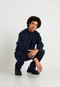 Mornarsko moder pulover s crewneck izrezom, sproščenega kroja, v kombinaciji s temnimi kavbojkami in črnimi robustnimi športnimi copati, z gladko teksturo in minimalnim dizajnom.