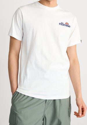Mann trägt ein weißes T-Shirt mit ellesse-Logo und graue Cargoshorts, steht mit einer Hand in der Tasche vor einem einfarbigen Hintergrund.