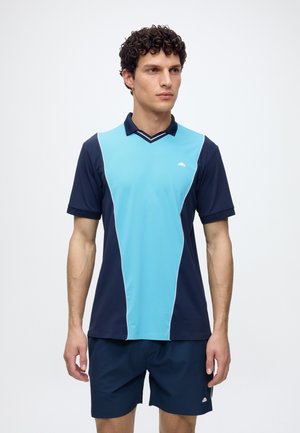 Homme portant un maillot de sport à manches courtes bleu marine et bleu clair et un short bleu marine, debout devant un fond blanc uni.