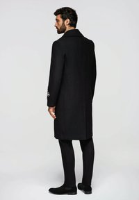 Schwarzer Wollmantel, mittellang, spitzer Kragen, Details mit seitlichen Taschen, glatte Textur. Kombiniert mit schwarzer Hose und Schuhen, das Modell von hinten gezeigt.