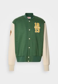Chaqueta varsity verde con mangas crema, con números amarillos bordados "1913" y "NEW YORK," y cierres a presión.