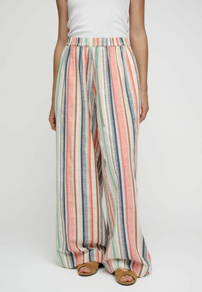 Femme portant un pantalon large à rayures verticales dans des couleurs pastel, associé à un haut blanc sans manches et des sandales marron.